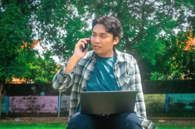 Bir kişi çimenli bir alanda oturuyor, dizüstü bilgisayar kullanıyor ve cep telefonuyla konuşuyor. Arka planda yemyeşil ağaçlar ve renkli bir duvar var. Bu da rahat ve verimli bir ortam olduğunu gösteriyor.