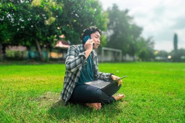 Parkta çimlerin üzerinde dizüstü bilgisayar kullanarak ve akıllı telefondan konuşarak oturan bir insan. Arka planda ağaçlar ve futbol hedefi yer alıyor. Bu da rahat bir açık hava çalışma ortamı olduğunu gösteriyor.