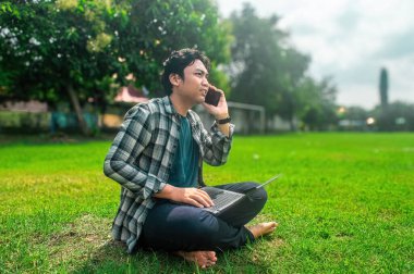 Çimenli bir alanda dizüstü bilgisayar kullanarak ve telefonla konuşarak bacak bacak üstüne atmış bir insan. Ağaçlar ve bir futbol golü rahat bir açık hava ortamı yaratır.