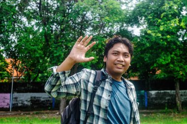 Ekose gömlek ve sırt çantası giyen biri, arka planda yemyeşil ağaçlar olan bir parkta el sallıyor.