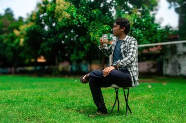 Parkta bir taburede oturan, kavanozdan kahve içen, ekose gömlek ve kot giyen, arka planda yemyeşil ağaçlar olan bir adam.