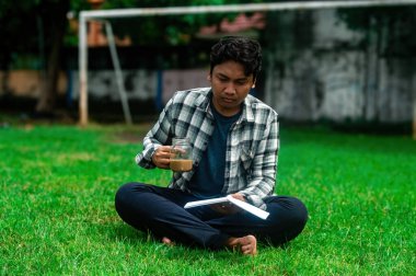 Ekose gömlekli ve siyah pantolonlu bir adam yeşil ağaçlı bir parkta elinde cam bir kupa kahve ve bir defter ile çimenlerin üzerinde bacak bacak üstüne atarak oturuyor.