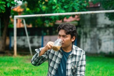 Ekose gömlekli bir adam arka planda futbol golü olan yeşil bir ortamda cam bardaktan kahve içer. Sahne tesadüfi ve rahatlatıcı bir anı yakalar.