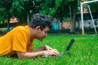 Turuncu gömlekli bir adam parkta çimlerin üzerinde dizüstü bilgisayar kullanıyor. Arka planda ağaçlar ve rahat bir açık hava ortamını gösteren bir futbol kalesi yer alıyor.