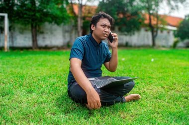 Bir insan parkta çimenlerde oturuyor, kulağına telefon dayayıp kucağında dizüstü bilgisayar kullanıyor, uzaktan kumandalı bir çalışma ya da açık hava ortamında çalışma öneriyor.