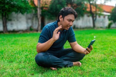 Adamın biri çimlerin üzerinde bağdaş kurmuş elinde akıllı bir telefonla video görüşmesi yapıyor. Arka planda ağaçlar ve duvarlar olan bir bahçe var.