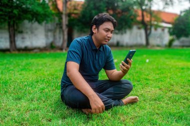 Adamın biri çimenli bir çimenlikte bacak bacak üstüne atmış, elinde akıllı bir telefonla oturmuş video görüşmesi yapıyormuş gibi görünüyor. Arka planda ağaçlar ve bir duvar var.