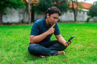 Çimlerin üzerinde bacak bacak üstüne atmış bir adam, elinde akıllı bir telefonla video görüşmesi yapıyormuş gibi görünüyor. Koyu mavi gömlek ve siyah pantolon giyiyor. Açık hava parkında düşünceli bir ifade takınıyor.