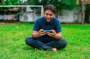 Adamın biri çimlerin üzerinde oturmuş, akıllı telefonuyla mobil oyun oynuyor. Dış mekanda bulanık bir kale direği ve ağaçlar var. Bu da rahat ve rahat bir ortam olduğunu gösteriyor.