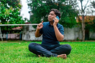 Bir adam parkta çimlerin üzerinde bacak bacak üstüne atmış akıllı bir telefonla konuşuyor. O koyu mavi bir gömlek ve siyah pantolon giyiyor, ağaçlar ve arka planda bir duvar var.