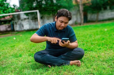 Adamın biri çimlerin üzerinde bacak bacak üstüne atmış iki eliyle akıllı telefon kullanıyor. Açık hava ortamında ağaçlar ve arka planda bir kale direği var. Bu da rahat ve rahat bir ortam olduğunu gösteriyor.