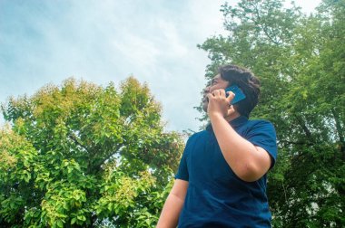 Mavi gömlekli bir adam açık havada, yemyeşil ağaçlar ve açık gökyüzü ile çevrili bir cep telefonuyla konuşuyor. Ayar, doğada gelişigüzel bir iletişim anını yakalıyor.