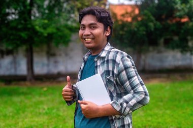 Bir öğrenci elinde bir defter tutuyor ve açık havada bir jest yapıyor. Ekose gömlek ve mavi tişört giyip arka planda ağaçlar olan çimenli bir alanda dikiliyor.