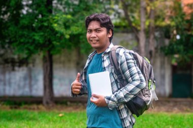 Elinde not defteri olan bir üniversite öğrencisi, bir parkta ekose gömlek ve mavi bir tişört giyerken başparmağını kaldırıyor. Arkaplanda yeşil çimenler ve ağaçlar yer alıyor