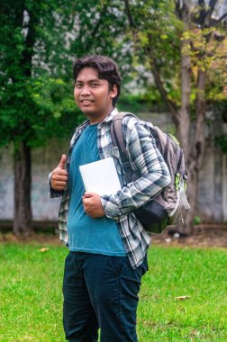 Bir üniversite öğrencisi elinde ekose gömlek ve sırt çantasıyla dışarıda duruyor, yeşil park geçmişi olan bir jest yapıyor.