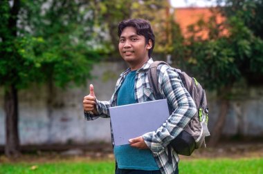 Bir üniversite öğrencisi dizüstü bilgisayarını tutuyor ve yeşil ağaçlar ve çimlerle çevrili dışarıda dikilirken baş parmak işareti yapıyor.
