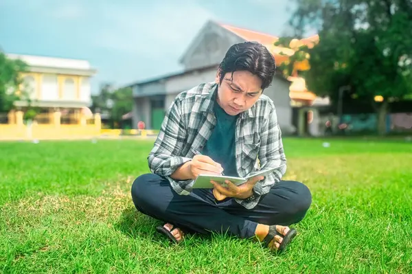Bir insan çimenli bir alanda bacak bacak üstüne atmış bir şekilde oturuyor ve bir deftere yazıyor. Arkaplanda binalar ve ağaçlar yer alıyor ve okumak veya günlük tutmak için ideal bir açık hava ortamı öneriliyor.