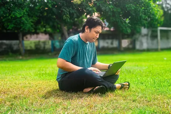 Çimenli bir arazide dizüstü bilgisayarla çalışan bir adam. Barışçıl açık hava manzarası ağaçlar ve güneş ışığını içeriyor, modern, uzak bir iş yaşam tarzını vurguluyor..