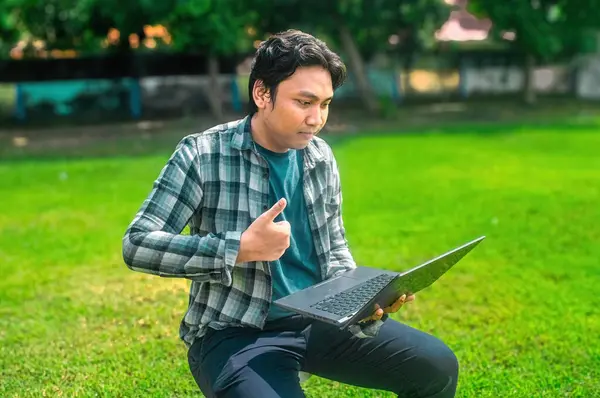 Parkta çimenlerde oturan, dizüstü bilgisayarını tutan ve baş parmak işareti yapan, ekose gömlek ve siyah pantolon giyen biri.