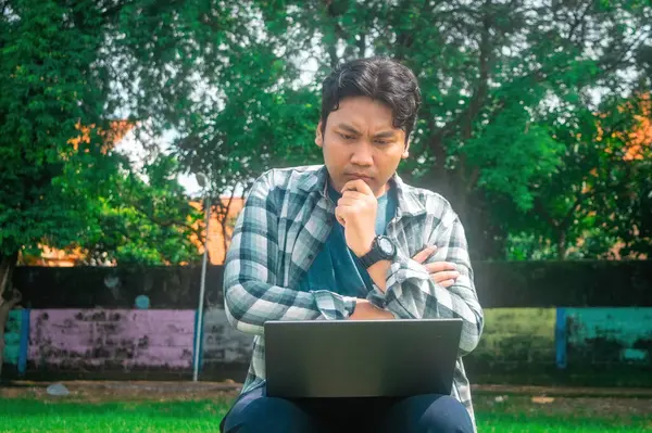 Çimenli bir alanda oturan, dizüstü bilgisayar kullanan, ekose gömlek ve kol saati takan, ağaçlı ve arka planda renkli bir duvar olan biri.