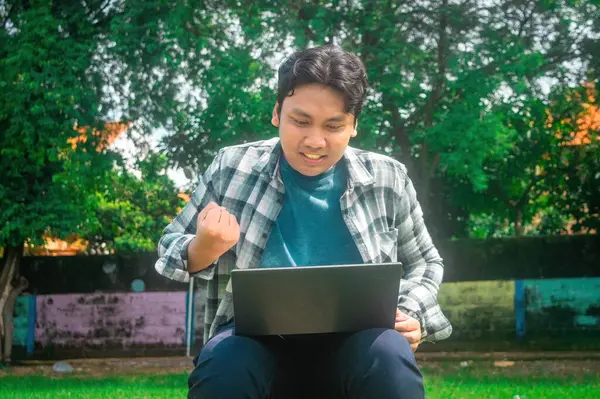 Dışarıda dizüstü bilgisayarla oturan, yumruk hareketi yapan, başarı ya da başarı duygusunu gösteren biri. Arkaplanda yemyeşil ağaçlar ve renkli bir duvar var.