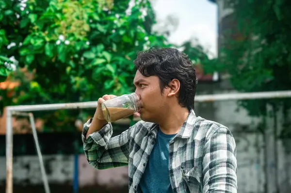 Ekose gömlekli bir adam arka planda yeşil yapraklar olan açık hava ortamında bir bardaktan kahve içer. Gündelik sahne bir rahatlama ve zevk anı yakalar.