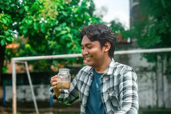 Ekose gömlekli bir adam dışarıda dururken bir kavanoz kahve tutuyor. Arka planda yemyeşil ağaçlar ve bulanık bir futbol kalesi var. Bu da rahat ve rahat bir ortam olduğunu gösteriyor.