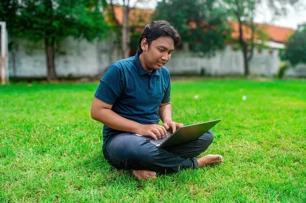 Adamın biri çimenlikte dizüstü bilgisayar kullanarak bacak bacak üstüne atmış oturuyor. Arka planda ağaçlar ve bulanık bir bina yer alıyor. Bu da uzak çalışma ve çalışma için ideal bir açık hava ortamı olduğunu gösteriyor.