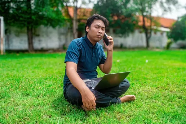 Parkta çimlerde oturan, dizüstü bilgisayar kullanan ve telefonla konuşan biri. Açık hava ortamında yeşillik ve ağaçlar sergileniyor, uzak bir çalışma ortamı veya çalışma ortamı sunuluyor