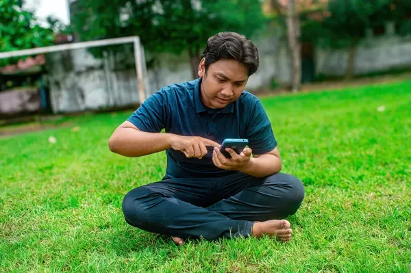 Adamın biri çimlerin üzerinde bacak bacak üstüne atmış iki eliyle akıllı telefon kullanıyor. Açık hava ortamında ağaçlar ve arka planda bir kale direği var. Bu da rahat ve rahat bir ortam olduğunu gösteriyor.