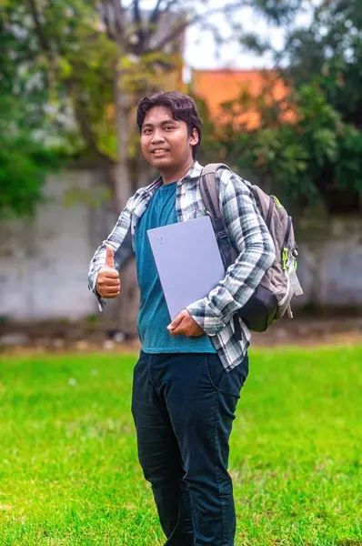 Çimenli bir alanda elinde dizüstü bilgisayarla başparmak kaldıran bir öğrenci. Öğrenci ekose gömlek, mavi tişört giyiyor ve sırt çantası taşıyor. Bu da rahat ve rahat bir atmosfer olduğunu gösteriyor.