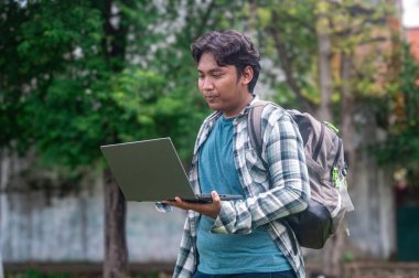 Bir üniversite öğrencisi dışarıda dizüstü bilgisayar ve sırt çantasıyla duruyor, ekose gömlek ve mavi tişört giyiyor, yeşilliklerle çevrili, çalışma ya da çalışma oturumunu öneriyor.