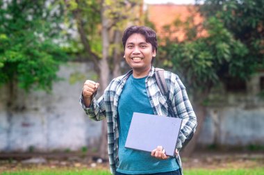 Ekose gömlekli ve mavi tişörtlü bir üniversite öğrencisi dışarıda dizüstü bilgisayarını tutuyor ve omzunda sırt çantası olan bir yumruk hareketi yapıyor.