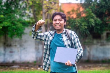 Ekose gömlekli ve sırt çantalı bir üniversite öğrencisi bir dizüstü bilgisayar tutuyor ve açık hava ortamında başarıyı ve kararlılığı simgeleyen bir yumruk kaldırıyor.