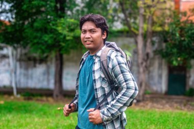 Ekose gömlek ve mavi tişört giyen bir üniversite öğrencisi dışarıda sırt çantasıyla bekliyor. Maceraya ya da okulda bir güne hazır. Arkaplanda yeşillik ve ağaçlar yer alıyor