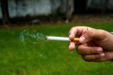 Yakın plan, yanan bir sigarayı tutarken havada dönen bir duman, bulanık yeşil bir arka plan üzerine kurulmuş. Bu resim sigara içme eylemini ve görsel etkisini vurgular.