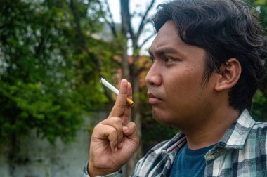 Bir insan açık havada iki parmağının arasında sigara tutuyor, ekose gömlek ve mavi tişört giyiyor. Arkaplanda doğal bir çevre oluşturan yeşillik ve ağaçlar yer alıyor.