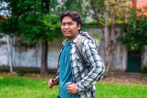 Ekose gömlek ve mavi tişört giyen bir üniversite öğrencisi dışarıda sırt çantasıyla bekliyor. Maceraya ya da okulda bir güne hazır. Arkaplanda yeşillik ve ağaçlar yer alıyor