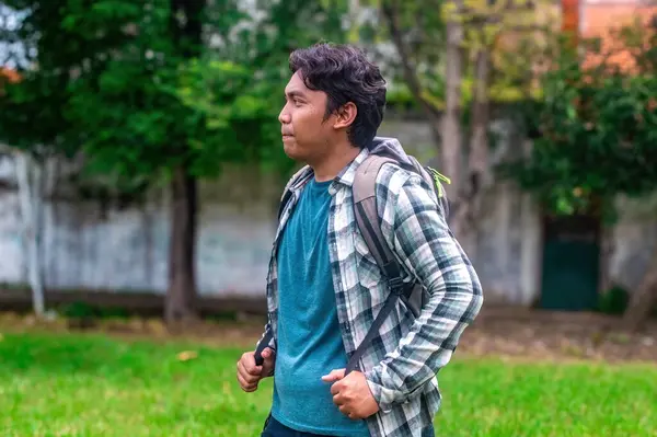 Mavi gömlek ve ekose ceket giymiş bir üniversite öğrencisi, sırt çantası taşıyor, dışarıda yeşillik ve arka planda bulanık bir duvarla duruyor. Okuldaki bir güne hazırım.