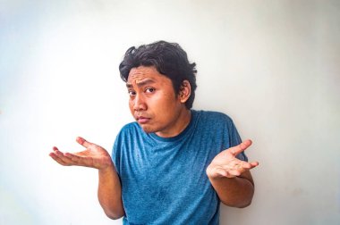 Mavi gömlekli bir adam düz bir zemine yaslanmış, elleri havada, avuçları havada, kafası karışmış ve şaşkın bir ifade sergiliyor.