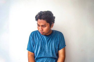 Mavi gömlekli bir adam düz beyaz bir arkaplanda oturuyor. Adamın kıvırcık saçları var ve aşağıya bakıyor, pişmanlık ya da tefekkür izlenimi veriyor.