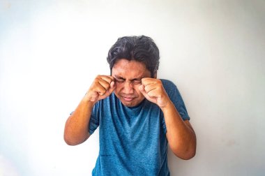 Mavi gömlekli bir adam ağlıyor gibi gözlerini ovuşturuyor. Arka plan bembeyaz, deneği açık seçik gösteriyor. Görüntü bir duygusal ifade anı yakalar.