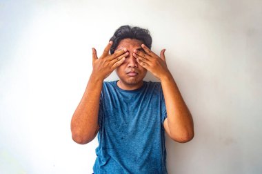 Mavi gömlekli bir adam iki eliyle düz beyaz bir arka plana sürtünüyor. Görüntü insani bir jest yakalıyor, muhtemelen yorgunluk ya da asabiyet belirtisi.