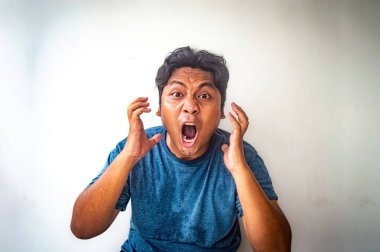 Mavi gömlekli bir adam şaşırmış ve kızgın bir ifadeyle yüzünü düz bir arkaplanda tutan bir adam görülüyor.