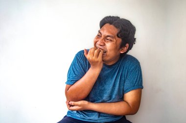 Mavi gömlekli bir adam düz beyaz bir arkaplanda oturuyor, diğer kolunu kucağında dinlendirirken parmağını ısırıyor. Bu resim tesadüfi ve samimi bir anı yakalar.