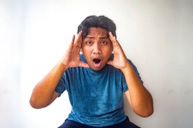 Mavi gömlekli bir adam ellerini yüzüne doğru tutuyor, şaşırmış ya da şok olmuş gibi görünüyor. Düz beyaz arka plan deneği açıkça öne çıkarıyor.