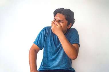 Mavi gömlekli bir adam düz beyaz bir zemine karşı oturuyor, burnunu eliyle kapatıyor, hoş olmayan bir şeyin kokusunu alıyormuş gibi. Görüntü, kötü kokuya karşı bir tepki anı yakalar.