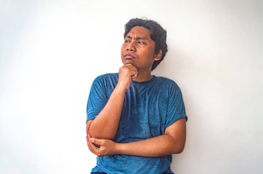 Mavi gömlekli bir adam beyaz bir arka plana karşı poz veriyor. Düşünceli bir yüz ifadesi var. Diğer kolu dirseğini desteklerken çenesini elinde dinlendiriyor.