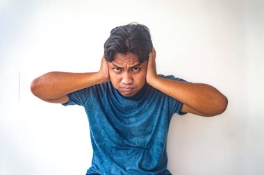 Mavi gömlekli bir adam muhtemelen gürültüyü engellemek için iki eliyle kulaklarını kapatıyor. Arka plan bembeyaz, adamın hareketlerini vurguluyor.