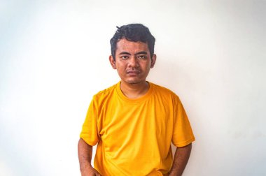 Sarı tişörtlü bir adam, tarafsız bir ifadeyle beyaz bir zemine yaslanır. Görüntü basit ve basit, sakin ve rahat bir pozu yakalıyor.